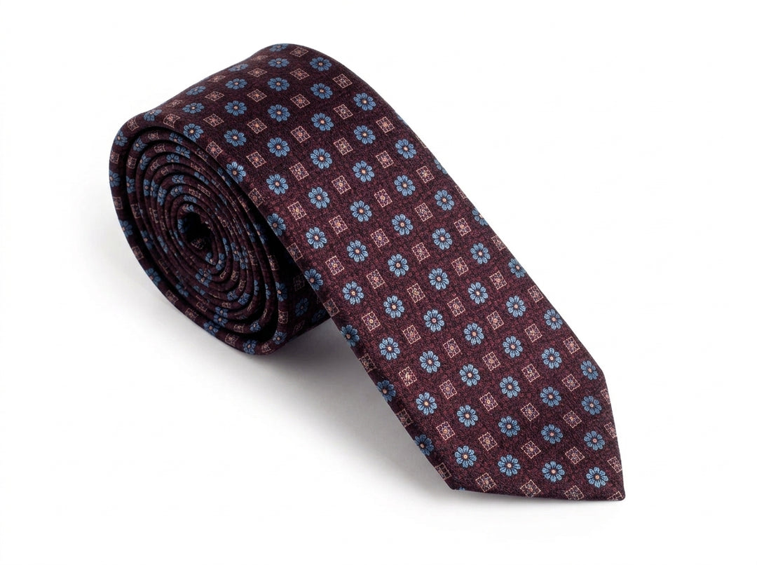 A rolled-up necktie featuring a repeating geometric pattern of blue and red motifs on a dark burgundy base, set against a plain white background.