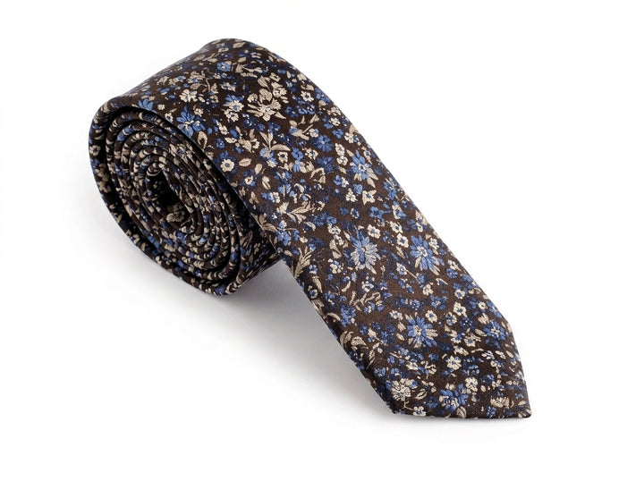 A rolled-up necktie featuring a dense floral pattern in blue, beige, and white on a dark brown background, set against a plain white studio backdrop.