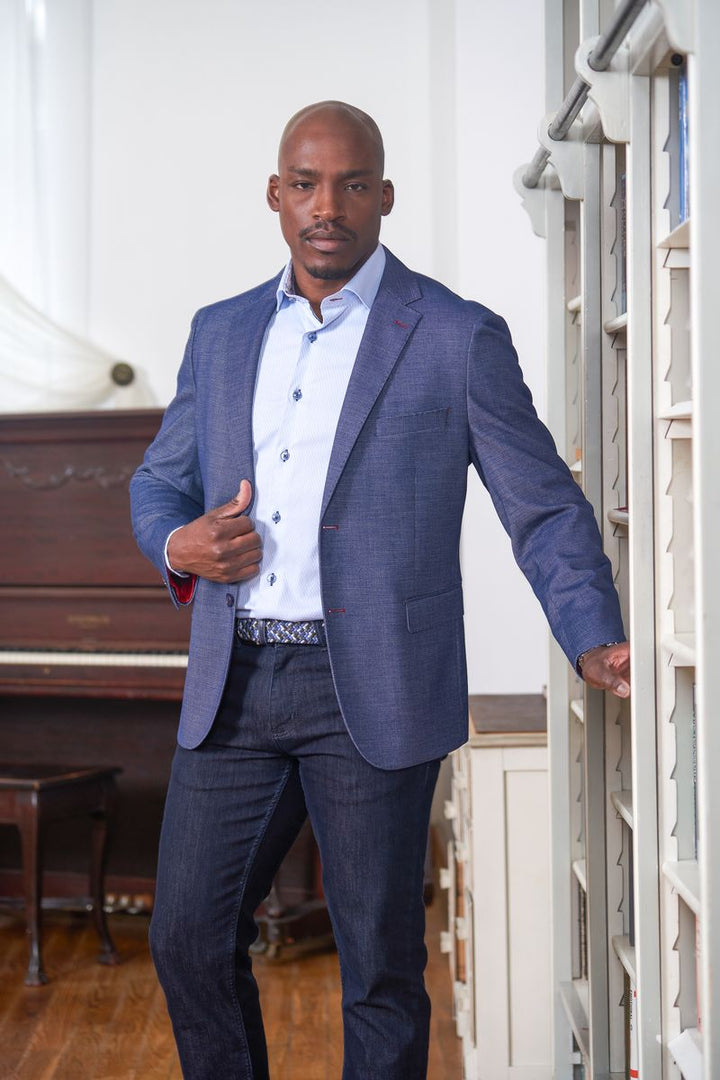Man in a blue suit standing in a room with a piano and bookshelf.