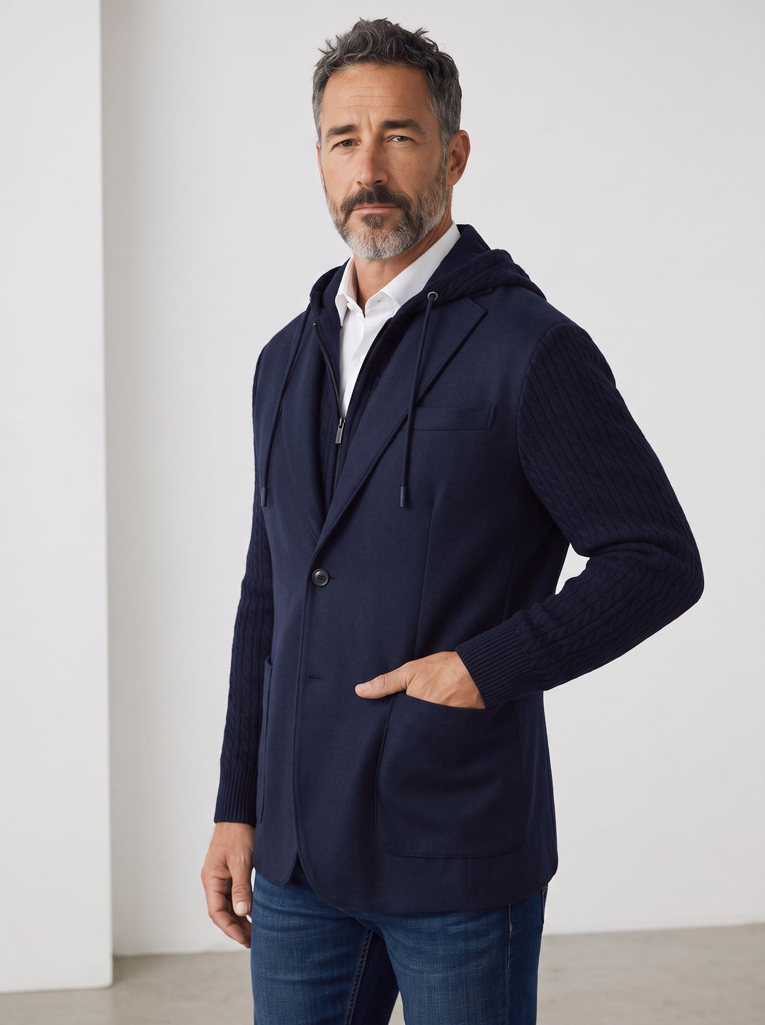 A man wearing a navy blue hooded zip-up blazer with ribbed sleeves, worn over a white collared shirt and blue jeans