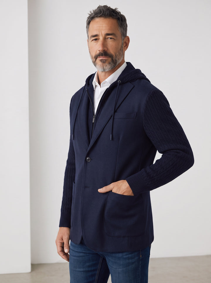 A man wearing a navy blue hooded zip-up blazer with ribbed sleeves, worn over a white collared shirt and blue jeans