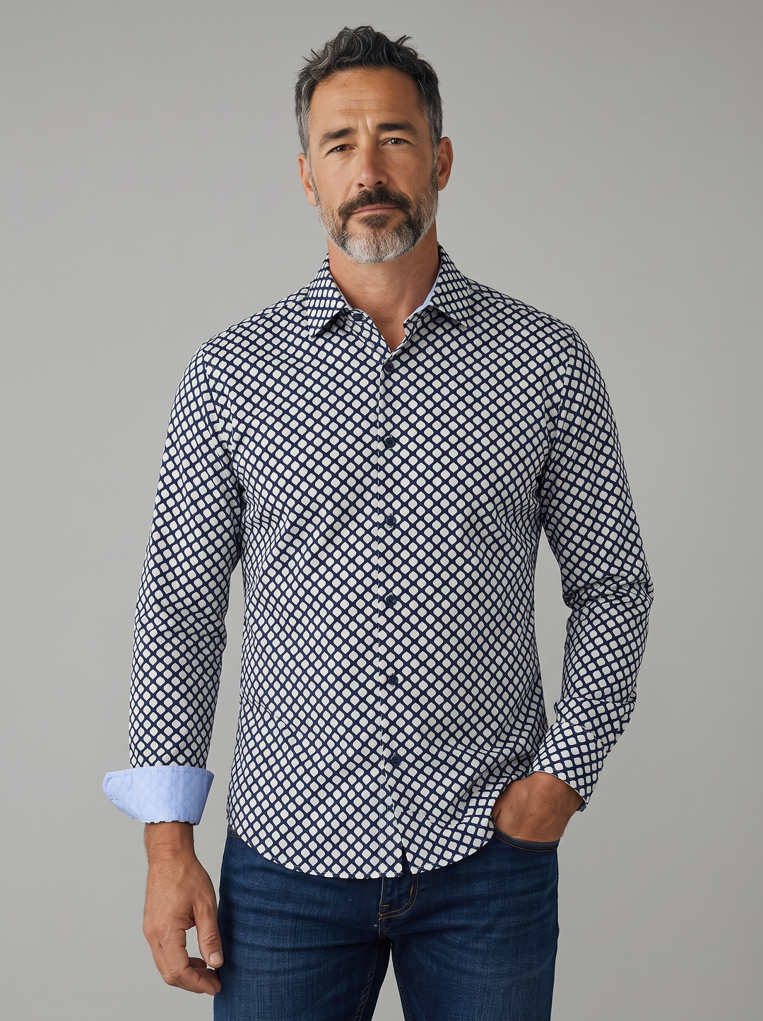 A man wearing a long-sleeve button-up shirt with a black and white geometric pattern, paired with dark blue jeans, standing against a solid gray studio background.