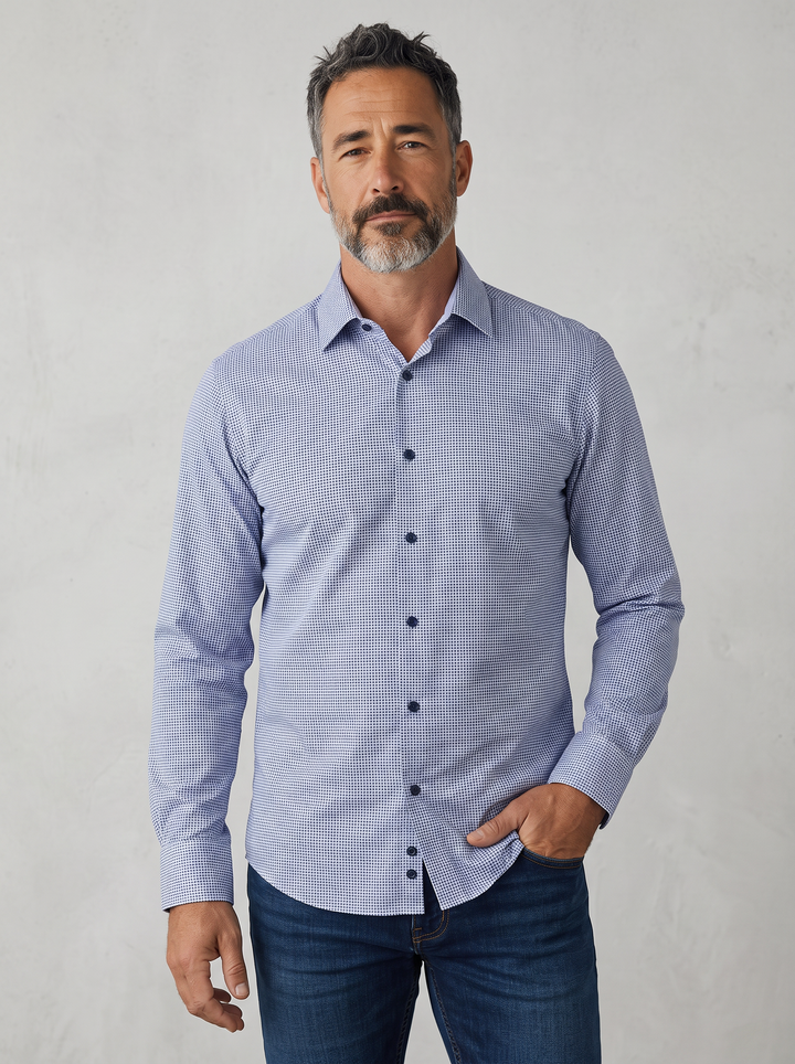 A man wearing a light blue long-sleeve button-down shirt with a subtle micro-check pattern and dark buttons, paired with dark blue jeans, standing against a textured light gray wall.