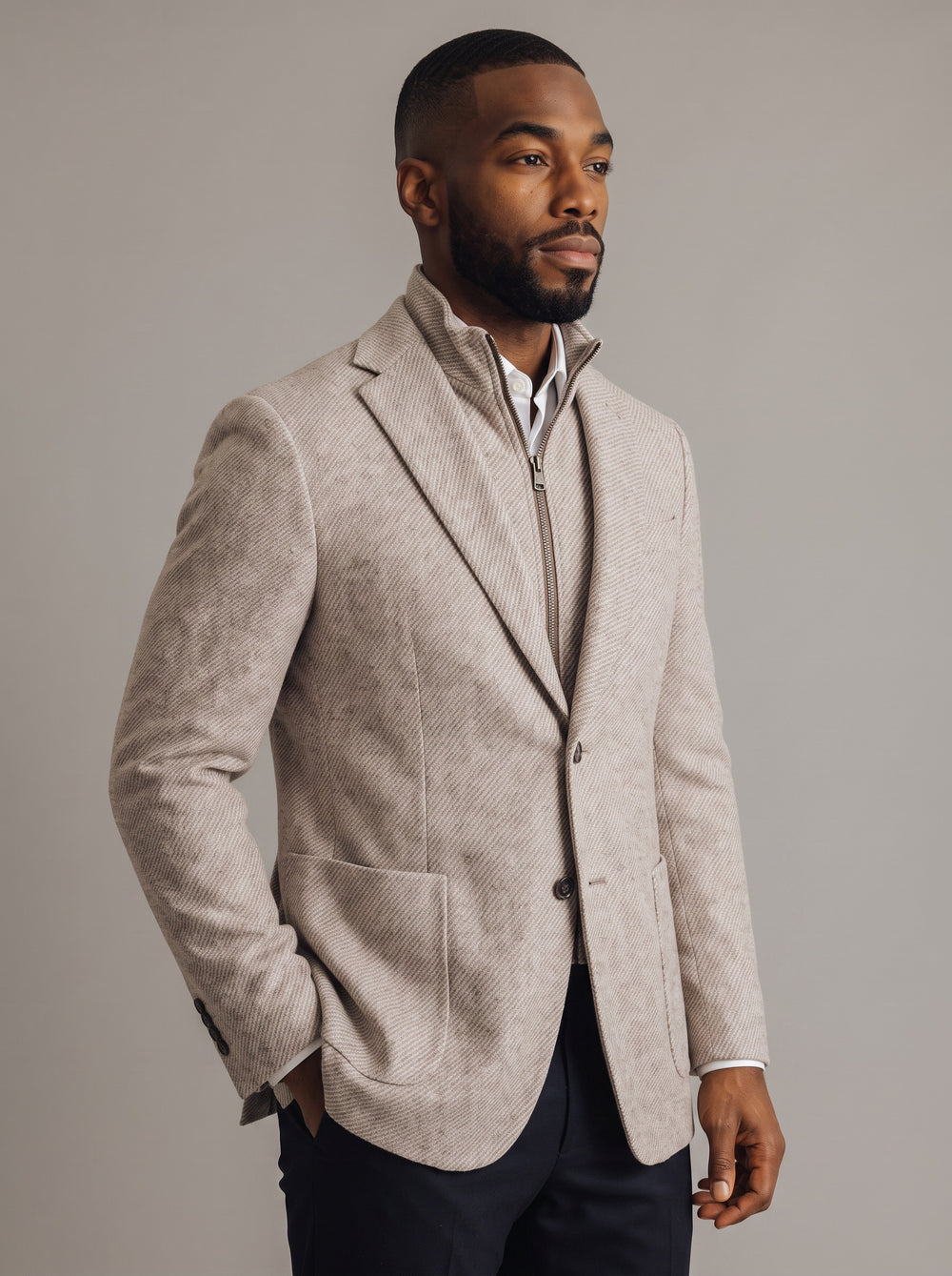 A man wearing a light beige textured blazer with a stand collar over a white shirt and dark trousers, photographed against a neutral gray studio background.