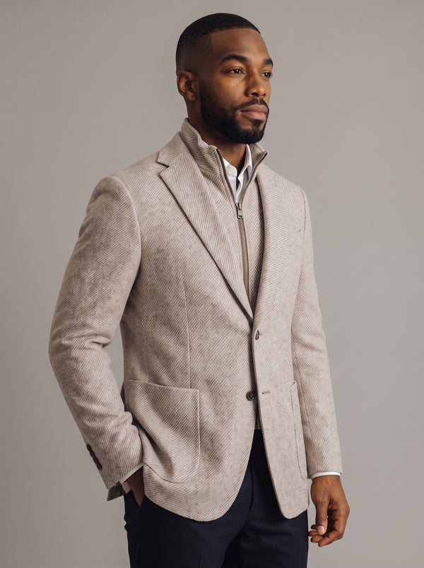 A man wearing a light beige textured blazer with a stand collar over a white shirt and dark trousers, photographed against a neutral gray studio background.