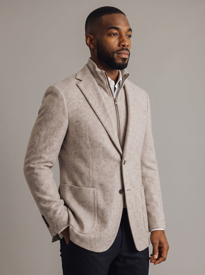 A man wearing a light beige textured blazer with a stand collar over a white shirt and dark trousers, photographed against a neutral gray studio background.