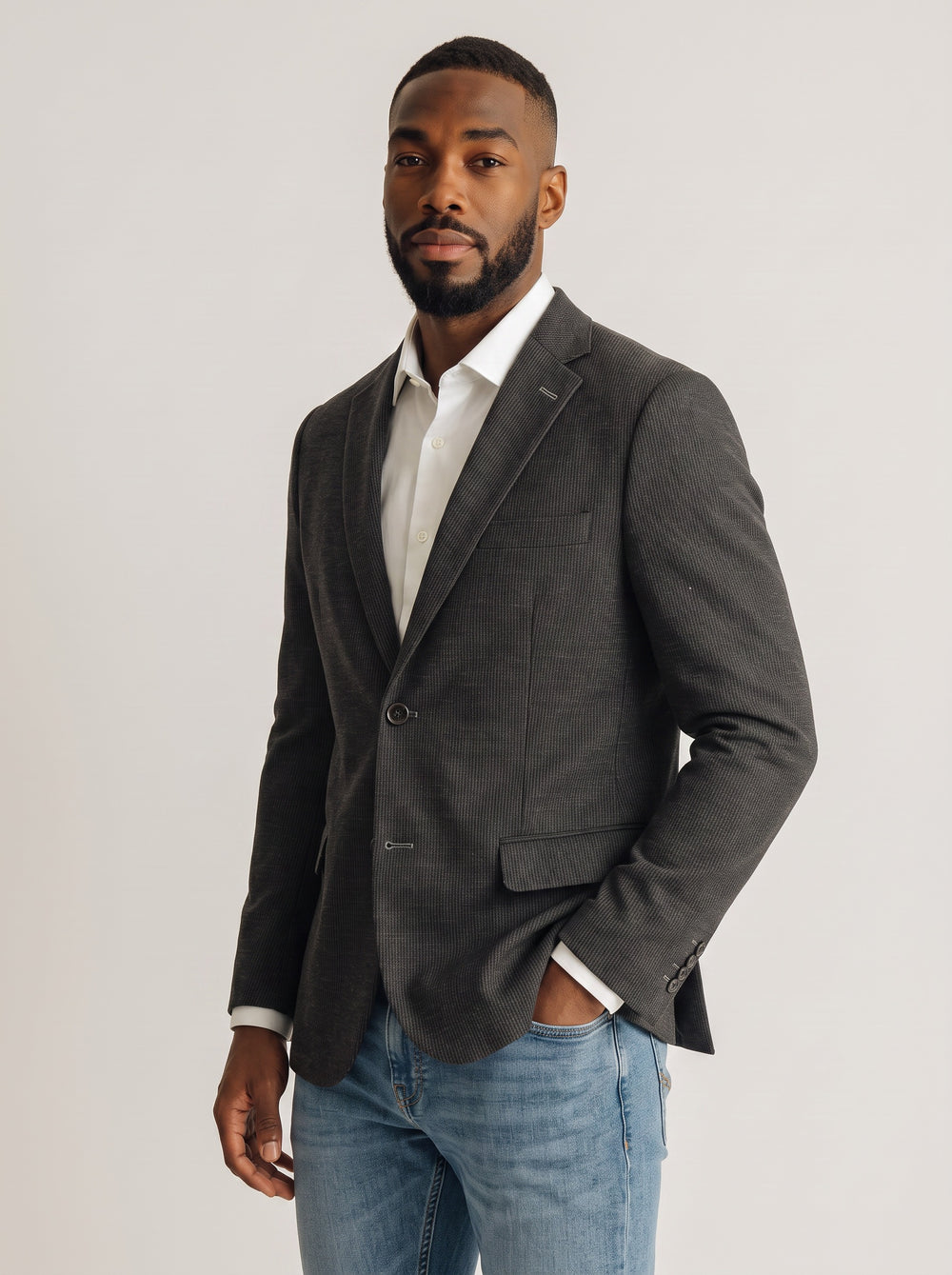 A man wearing a dark gray textured blazer over a white collared shirt and blue jeans, standing against a light gray studio background.