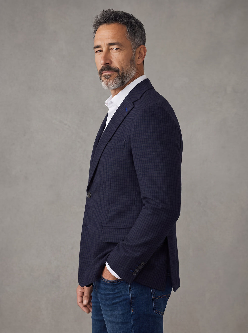 A man wearing a dark navy blue textured blazer over a white collared shirt and blue jeans