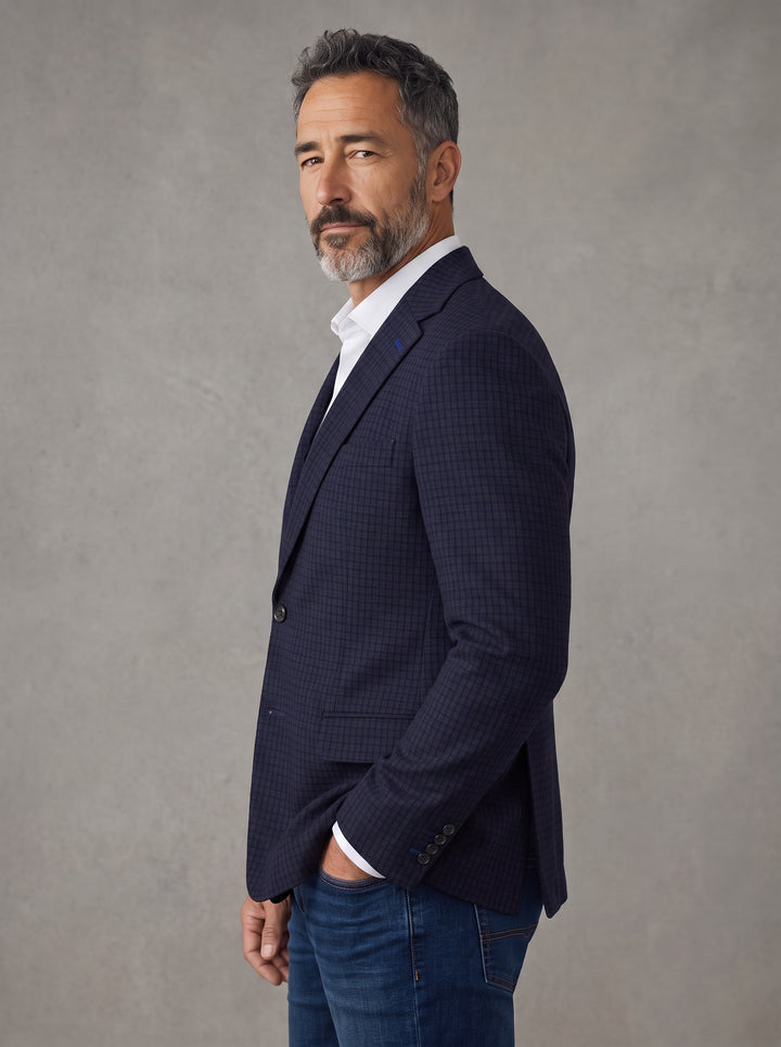 A man wearing a dark navy blue textured blazer over a white collared shirt and blue jeans