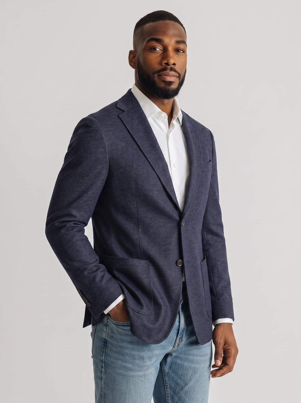 A man wearing a navy blue textured blazer over a white collared shirt and light blue jeans.