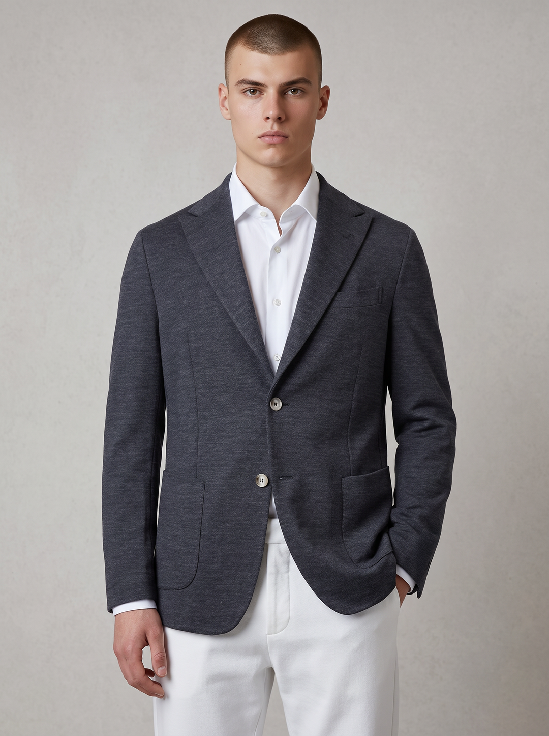 A male model wearing a dark gray heathered blazer over a white dress shirt and white trousers