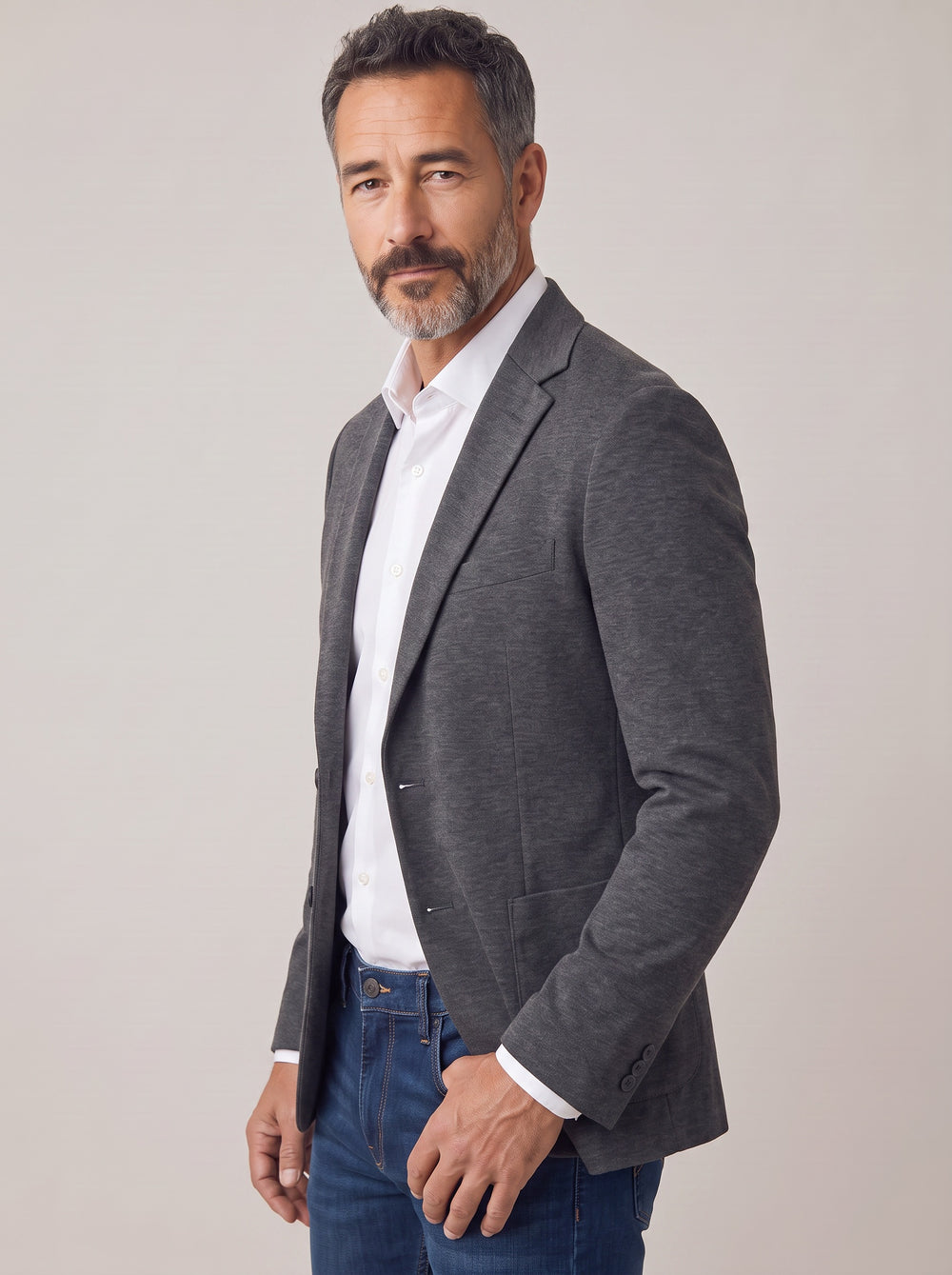 A man wearing a dark gray heathered blazer over a white collared shirt and blue jeans.