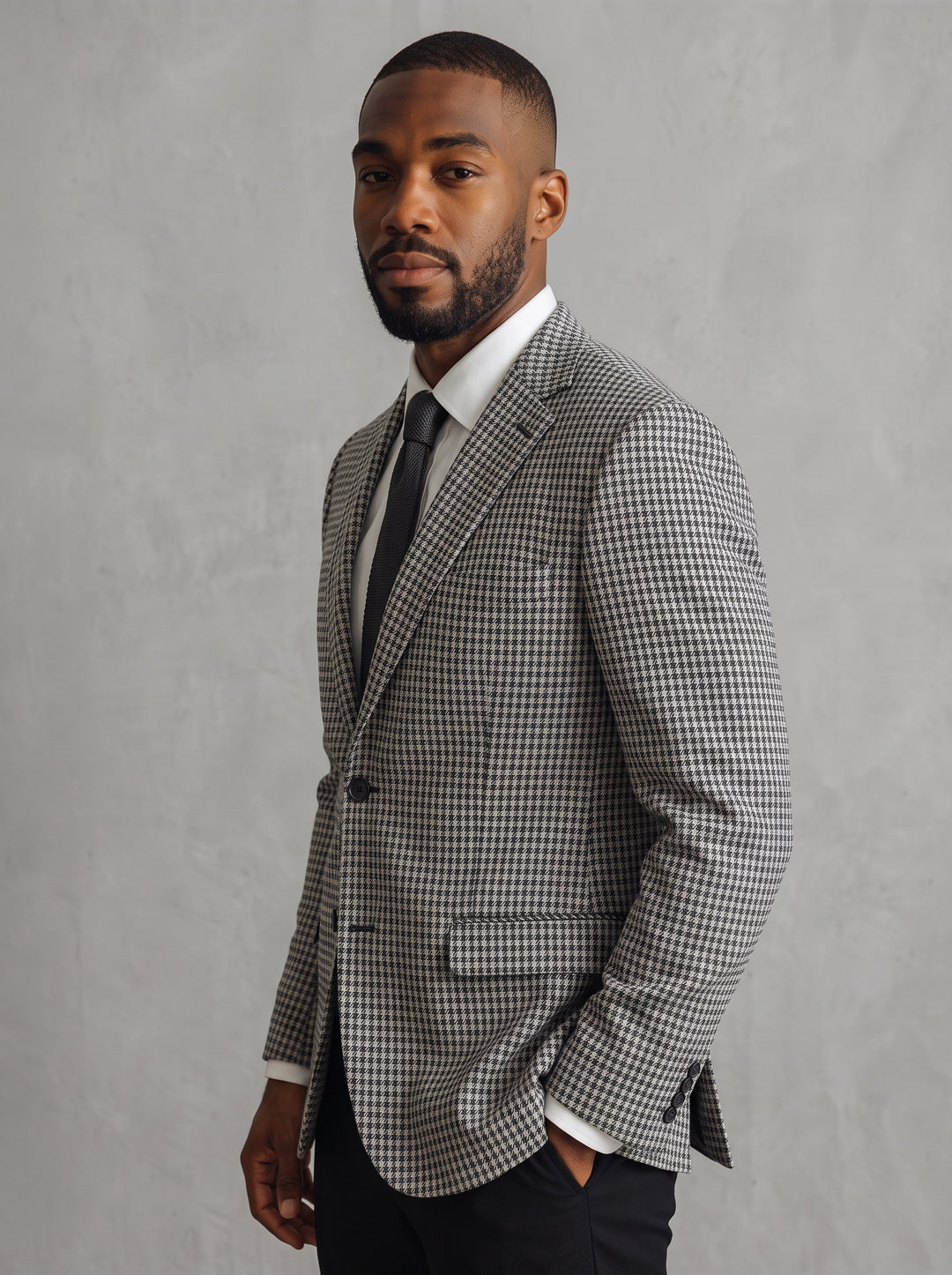 A man wearing a grey and white checkered blazer, white dress shirt, and black tie, standing against a textured gray background.