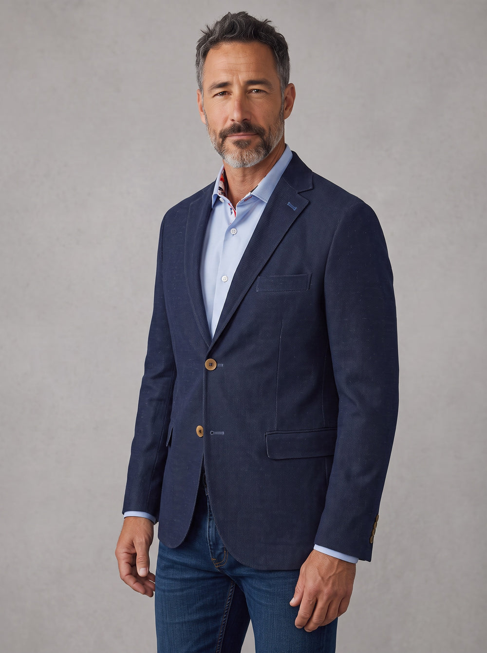 A man wearing a navy blue blazer over a light blue dress shirt and blue jeans,