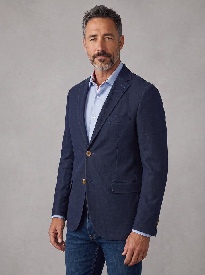 A man wearing a navy blue blazer over a light blue dress shirt and blue jeans,