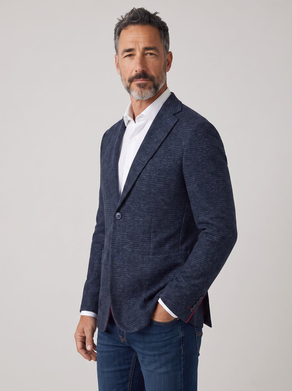A man wearing a textured navy blue blazer over a white collared shirt and blue jeans, standing against a light gray studio background.