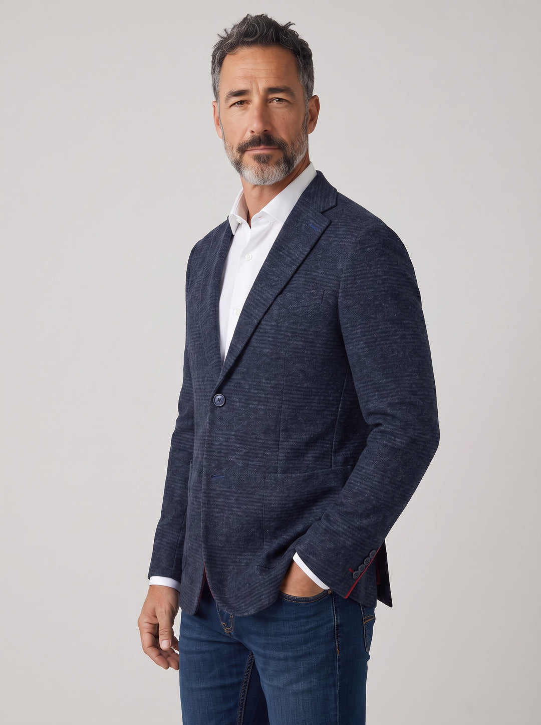 A man wearing a textured navy blue blazer over a white collared shirt and blue jeans, standing against a light gray studio background.