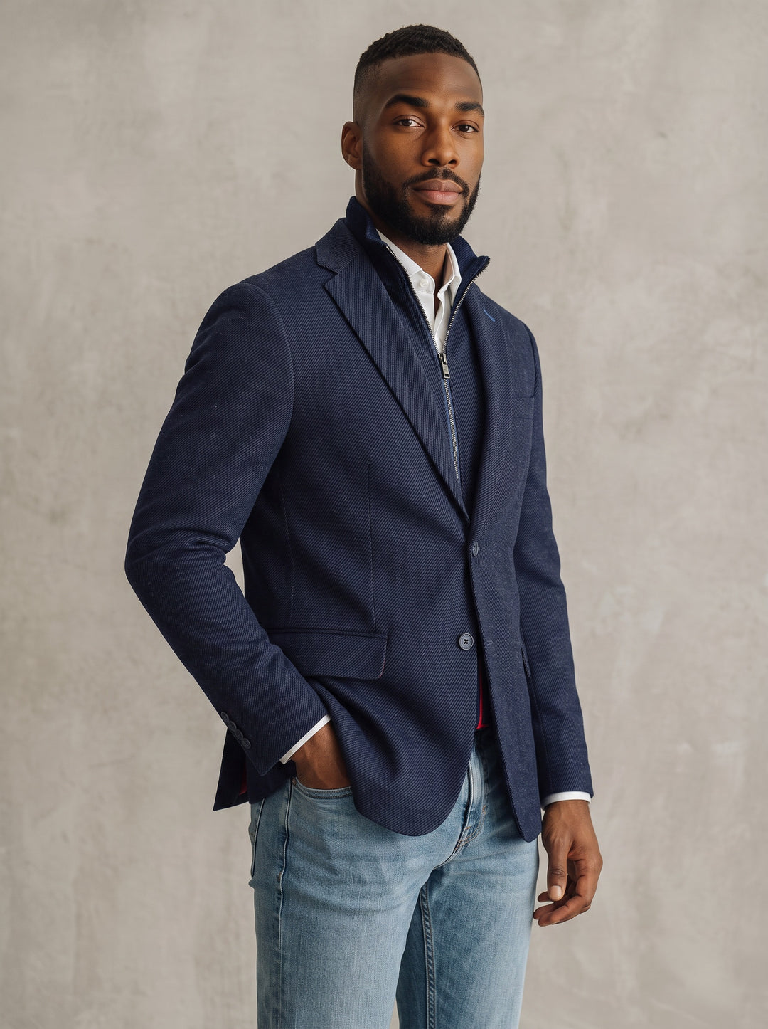 A man wearing a navy blue pinstripe blazer over a white zip-up shirt and light blue jeans.