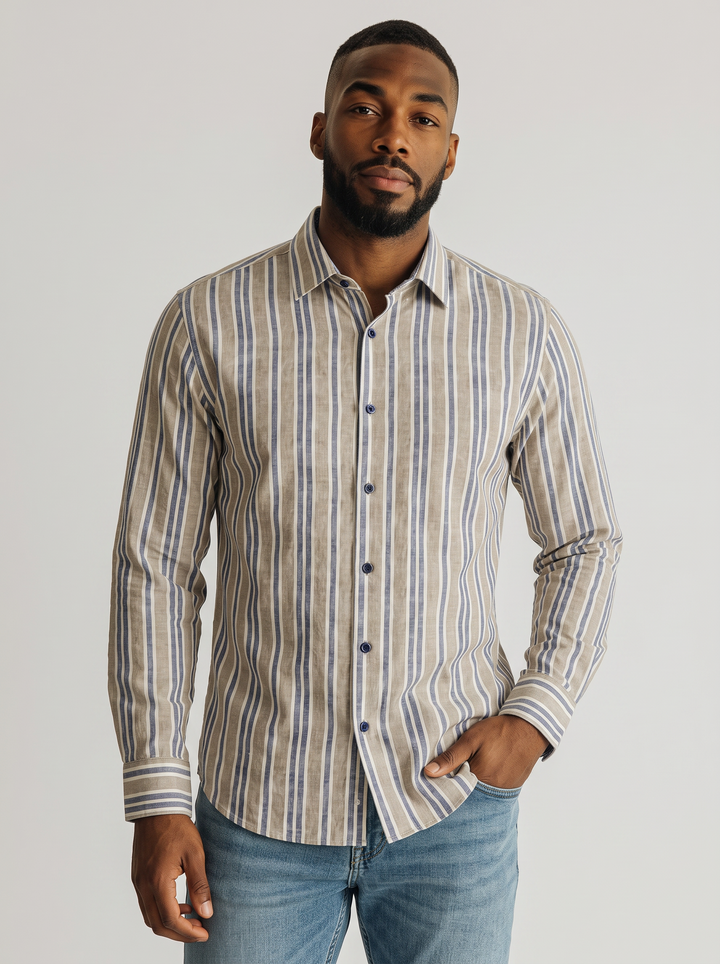 A man wearing a long-sleeve button-up shirt with vertical beige, brown, and blue stripes, paired with blue jeans, photographed against a light gray studio background.