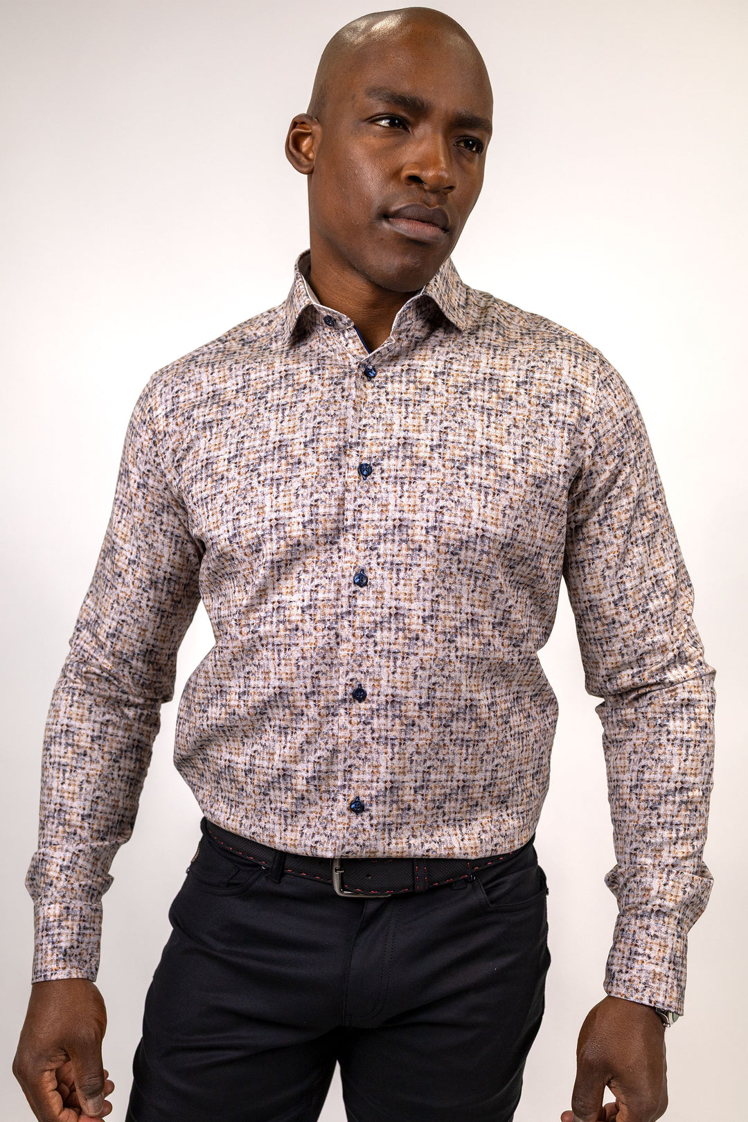 A man wearing a long-sleeve button-down shirt with a dense multicolor micro-pattern of beige, blue, and brown, paired with dark trousers, photographed against a light studio background.