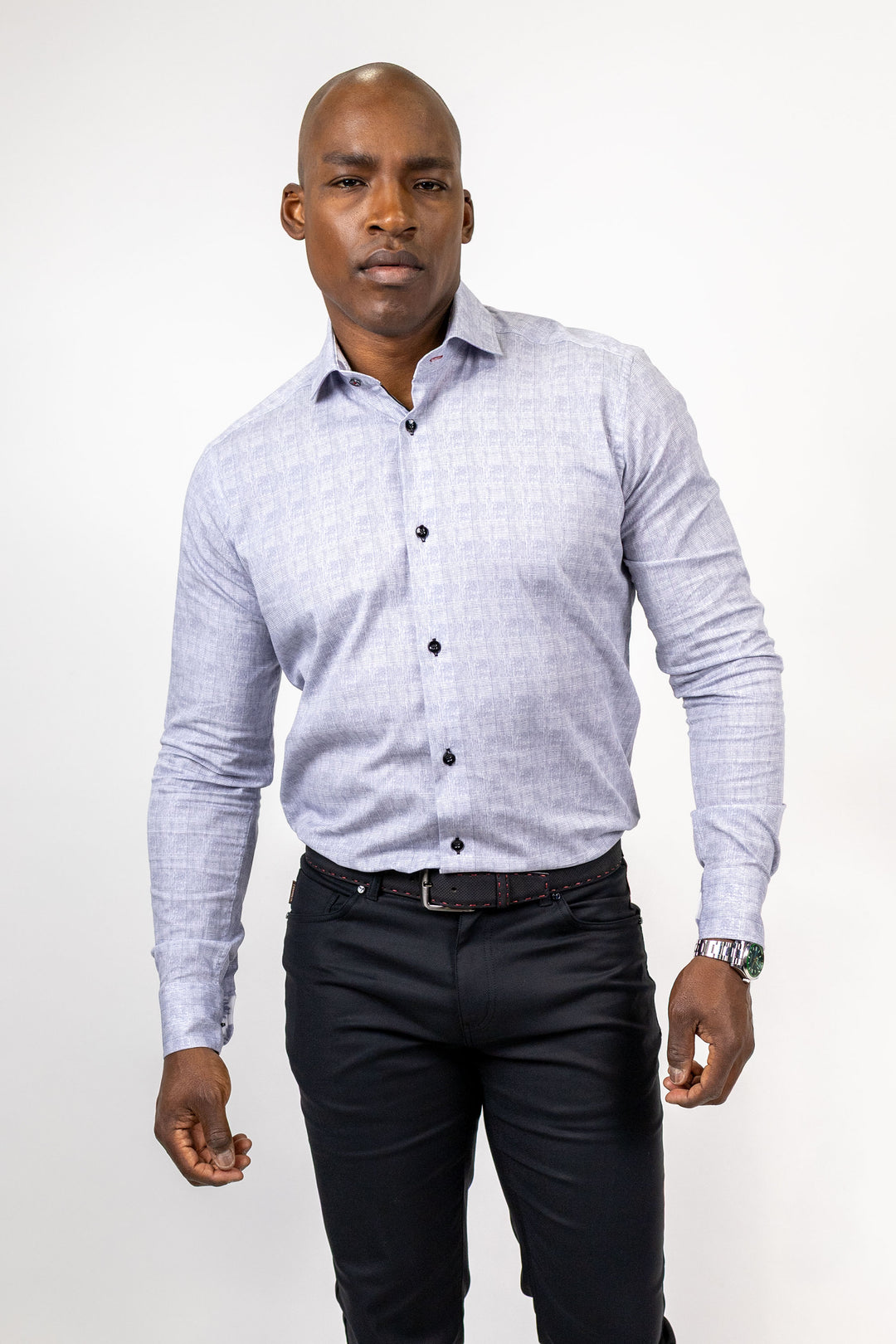 A man wearing a light blue and white patterned long-sleeve button-down shirt, paired with dark trousers and a black belt, standing against a plain white studio background.