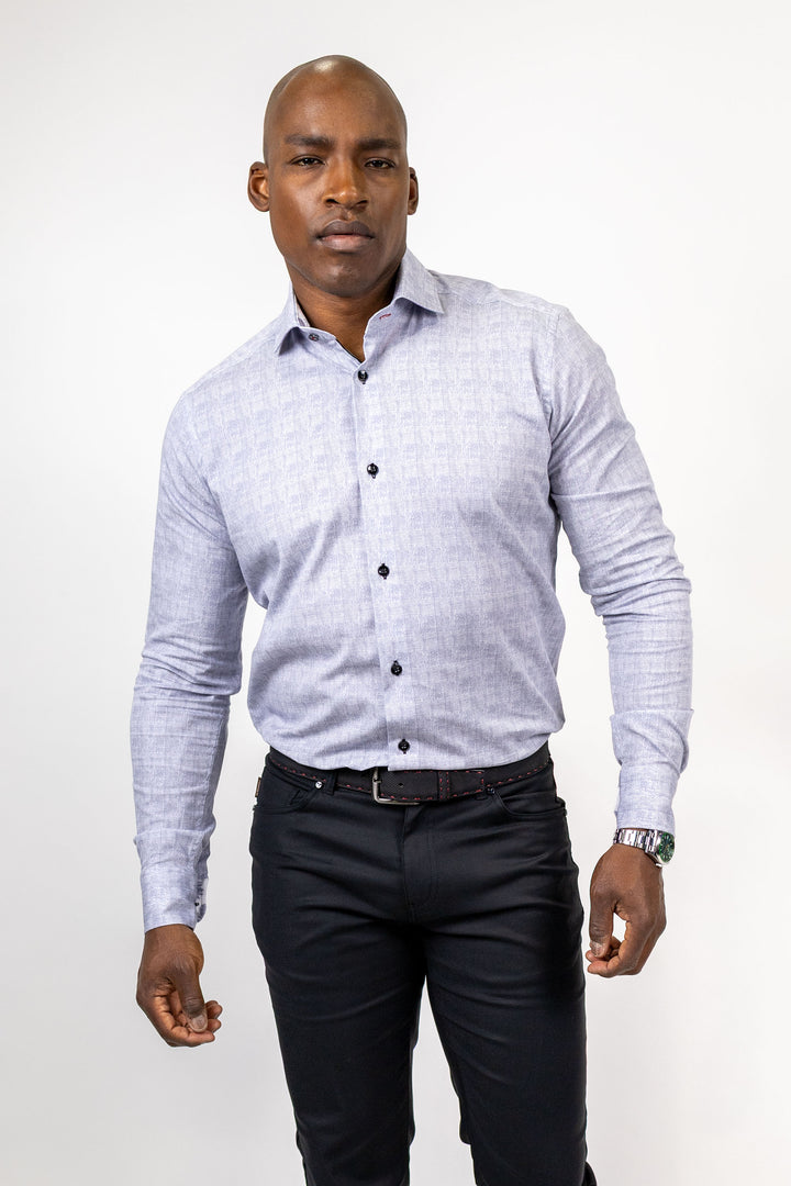 A man wearing a light blue and white patterned long-sleeve button-down shirt, paired with dark trousers and a black belt, standing against a plain white studio background.
